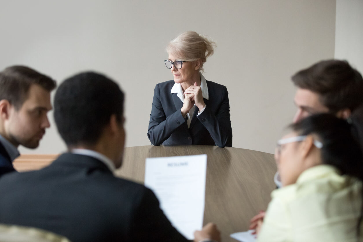 An older woman in a business suit sits pensive at a meeting table with younger colleagues, symbolizing age discrimination in the workplace and the need for legal support.